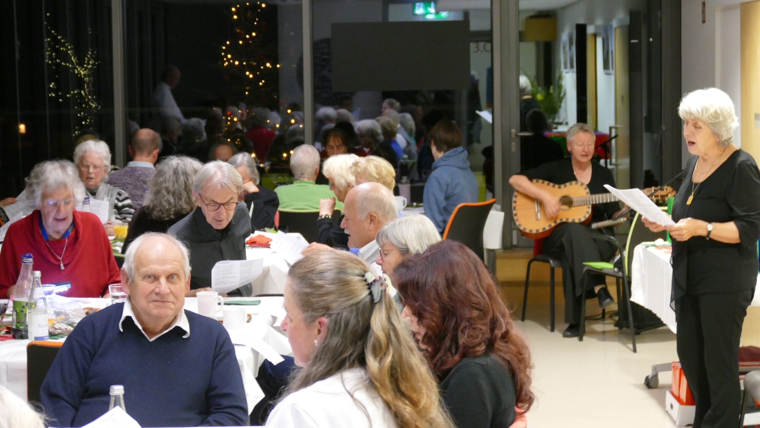 singen1 Mehrere Personen sitzen an Tischen in einem Raum, im Hintergrund spielt eine Person Gitarre und eine andere hält ein Blatt Papier in der Hand